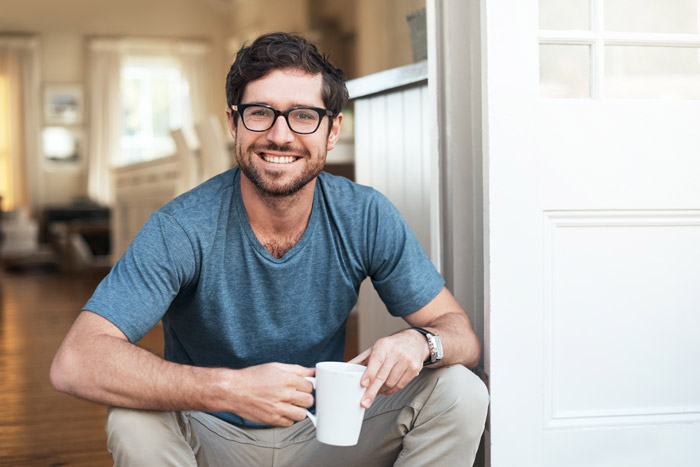 younger, thin man with dark hair and glasses holding a cup of coffee while sitting down and smiling