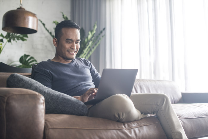 asian/pacific-islander male wearing a long sleeved blue shirt and khakis on a leather couch using his laptop